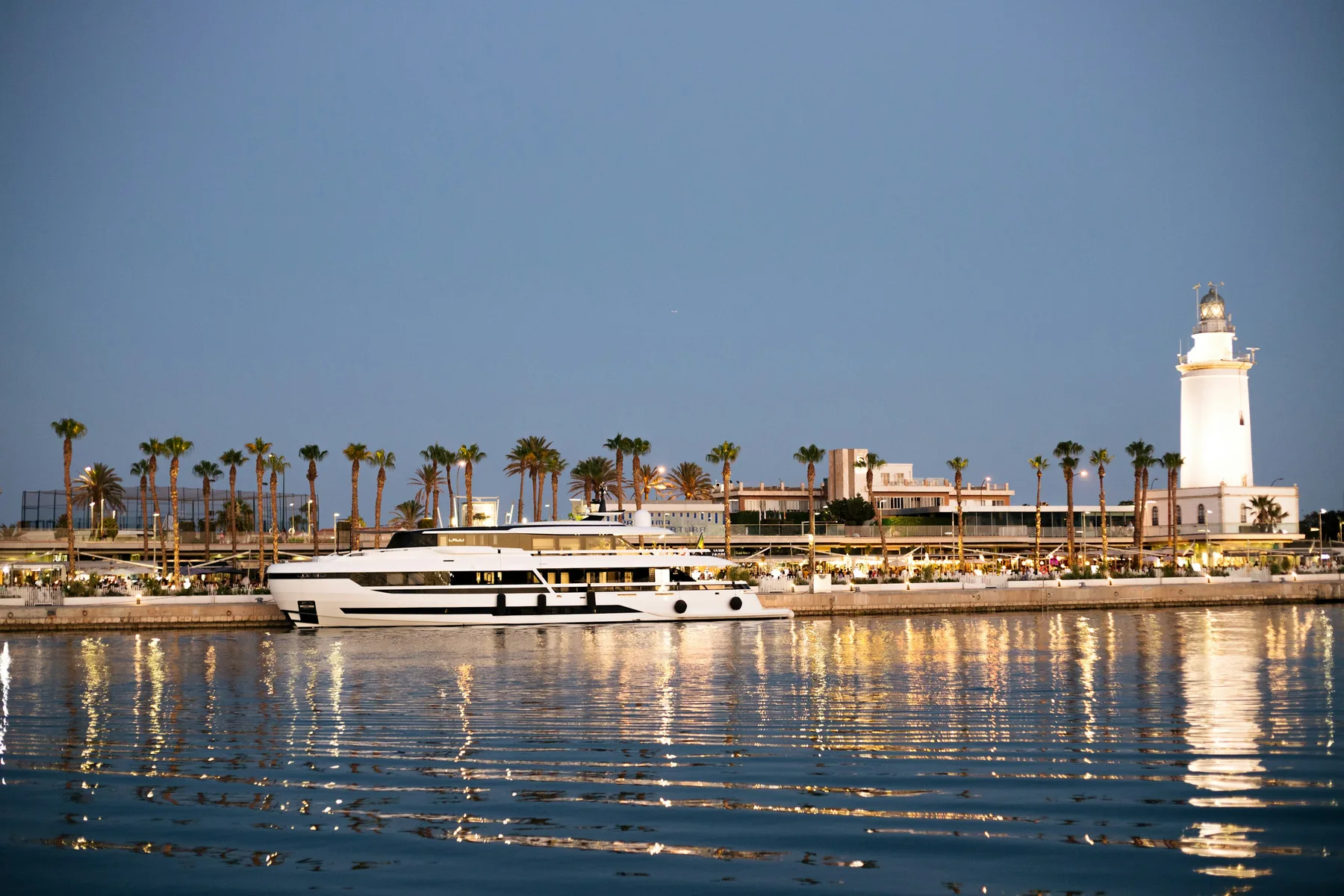 The white lighthouse and palm trees of the Málaga port at dusk, with a yacht moored on calm water